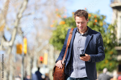 Young urban businessman professional on smartphone walking in street using mobile phone app texting sms message on smartphone wearing smart casual jacket. City lifestyle commute person walking.