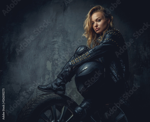 Blonde female in leather clothes sits on a car's wheel .