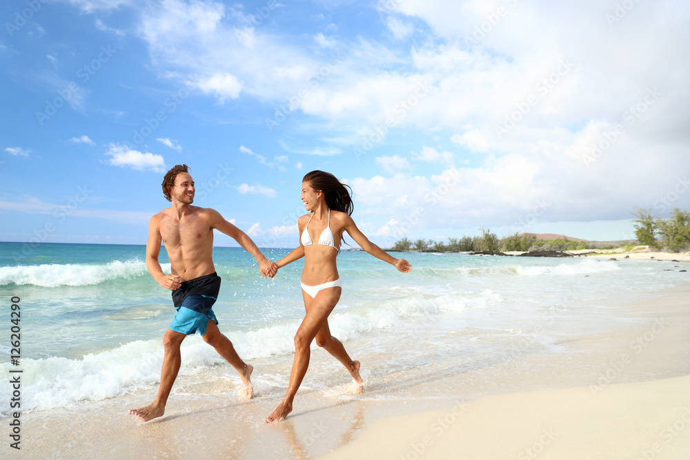 Beach summer vacation couple running on holidays. Happy fun beach vacations couple walking together laughing having fun on travel destination. Playful interracial couple in swimwear holding hands.