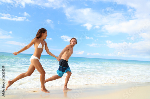 Happy beach couple vacation getaway. Young people in bikini and swimwear running holding hands together having fun on tropical beach paradise. Perfect blue ocean water and white sand.