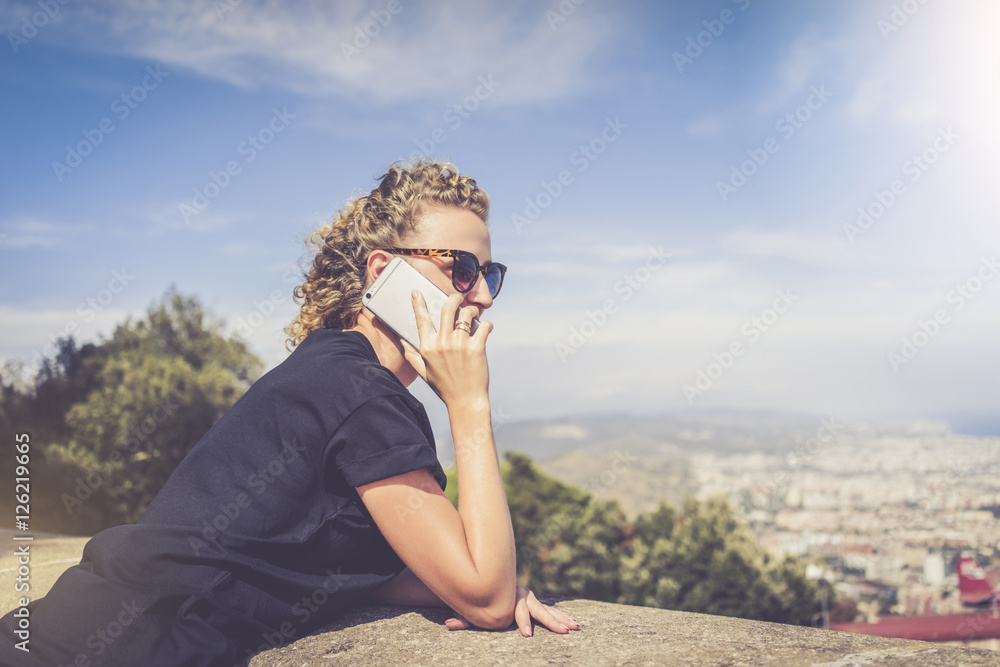 Side view of a young woman dressed in a black T-shirt and sunglasses, standing and talking on the phone. In the background landscape with aerial view, clouds in the sky. Girl uses the gadget.