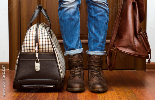 Девушка в джинсах и кожаных ботинках с рюкзаком и чемоданом стоит у двери