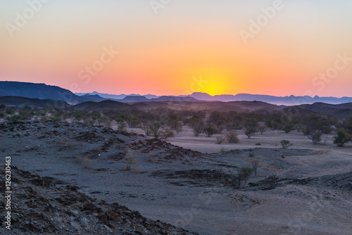 Colorful sunset over the Namib desert  Namibia  Africa. Mountains  dunes and Acacia trees silhouette in backlight. Orange red clear sky at the horizon.