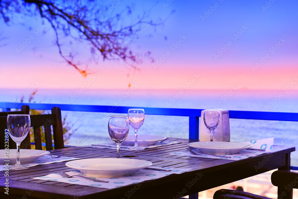 served table on the sea shore. glasses, plates, coasters saucer on brown wooden table. In the background, the sea and the light of the setting sun, sunset. Pleasant warm evening, 
