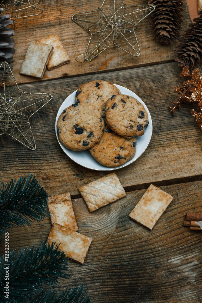 Chocolate cookies in a white plate on wooden background. Near cones and cinnamon stick, roughly. View from above. The theme of the new year holiday Chocolate cookies in a white plate on wooden background. Near cones and cinnamon stick, roughly. View from above. The theme of the new year holiday