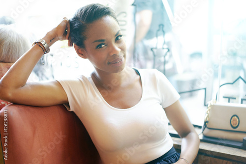 A beautiful african woman is looking at the camera while sitting in a city coffee shop during brunch time. A waist up photo of a female freelancer having rest in a cafe on a blurred background.