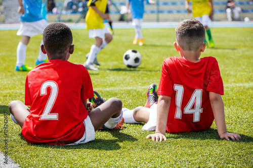 Children football soccer tournament. Kids playing football match. Two boys friends caucasian and african in red shirts watching soccer game competition. 
