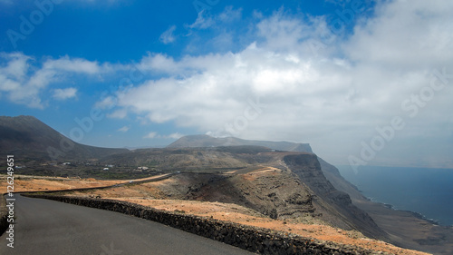 Asphalt road over a cliff above the ocean disappearing over the horizon through volcano mountain hillsides. White clouds on a blue sky. Lanzarote, Canary Islands, Spain