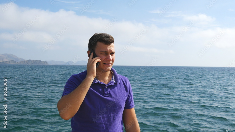 Young handsome man is talking on mobile phone on a sea beach. Serious guy speaking on the cellphone on the background of the ocean. Call from the summer vacation. Beautiful landscape. Close up