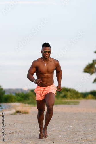 Young  beautiful  athletic muscular black guy running along sandy beach. Man fitness model with a beautiful body.