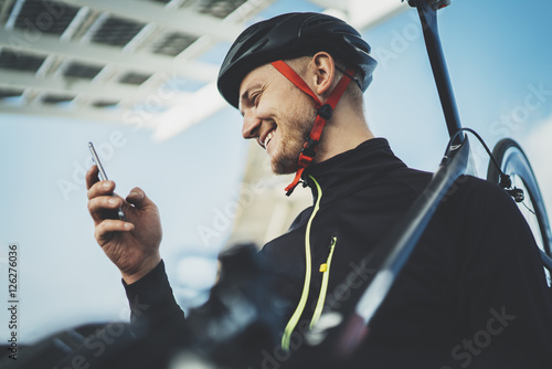 Young happy smiling cyclist man using smartphone while resting after workout, road bicycle on arms, cheerful sportsman checking fitness results via cellphone while riding a bike at the sunny morning