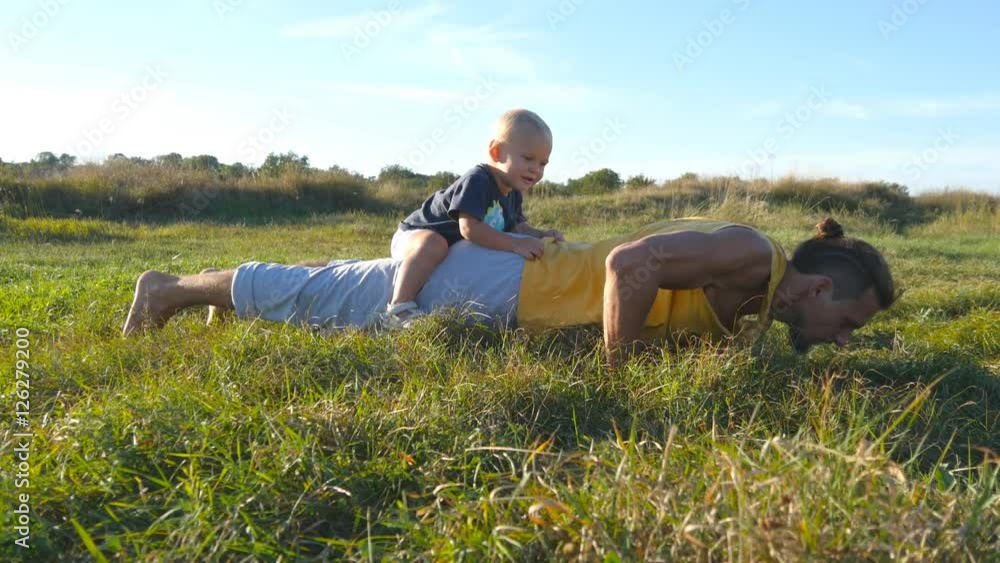 Young muscular father doing push ups with his little son on his back. Athletic man doing push-ups at green grass with happy smiling baby sitting on his rear. Strong guy spending time with your child Young muscular father doing push ups with his little son on his back. Athletic man doing push-ups at green grass with happy smiling baby sitting on his rear. Strong guy spending time with your child