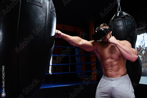 Strong boxer fights in VR glasses/Muscular boxer with glasses of virtual reality hit black punching bag in dark gym.Selective focus