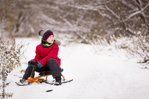 Portrait of cute kid boy sitting on sledge on the snow on a winter day. Child playing outdoors. Lifestyle concept