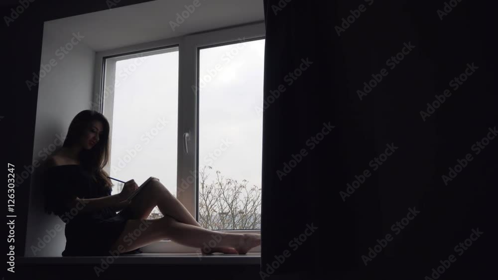 Attractive young brunette girl sitting on the windowsill and writing in notebook Attractive young brunette girl sitting on the windowsill and writing in notebook
