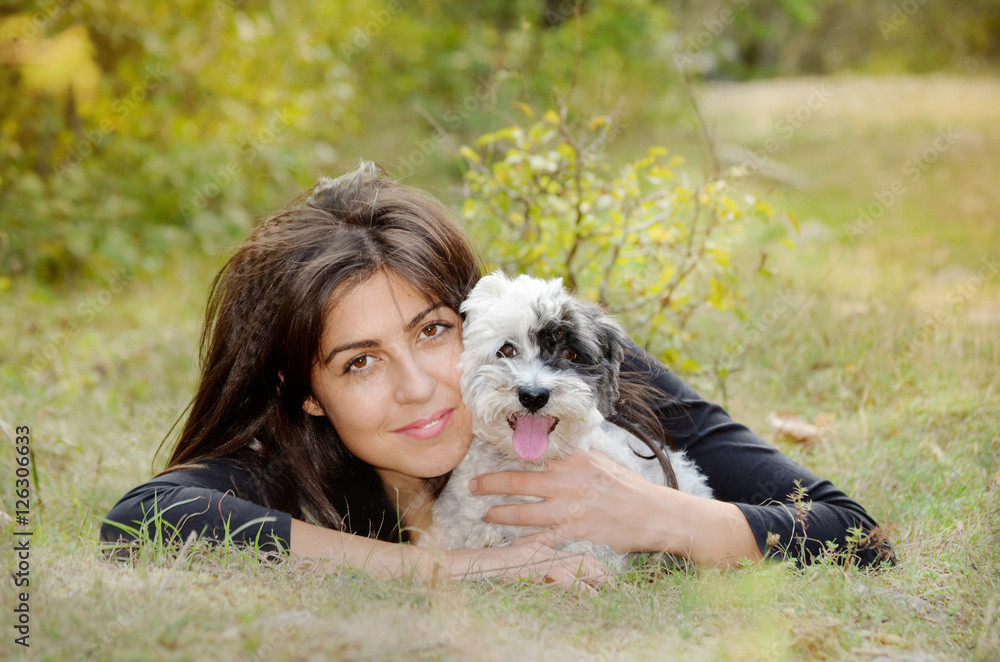 smiling woman laying on a meadow with her small havanese dog