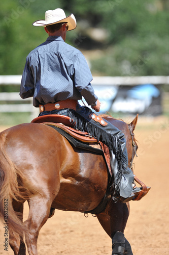The rear view of a rider in cowboy chaps, boots and hat on a horseback running ahead and stopping the horse in the dust.