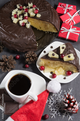 Homemade chocolate-glazed pineapple cake with one slice on white plate and a cup of coffee with three pieces of chocolate on grey background with christmas decorations