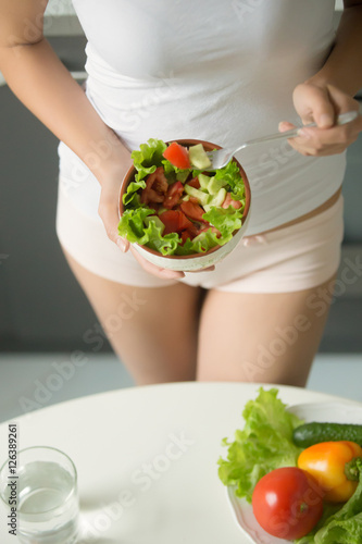 Female wearing underwear panties and white color tank top holding a bowl of green salad against her waist near the kitchen table, preparing to it with a fork, close up. Vertical, weight loss concept