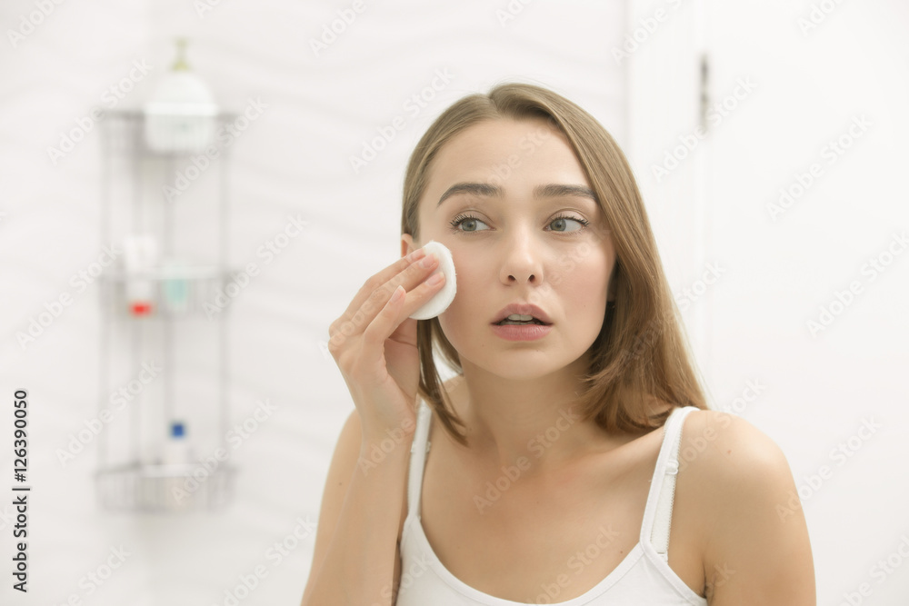 Focused beautiful young woman cleaning her skin with a cotton pad, looking at the mirror at home bathroom. Beauty, skin care concept, lifestyle Focused beautiful young woman cleaning her skin with a cotton pad, looking at the mirror at home bathroom. Beauty, skin care concept, lifestyle