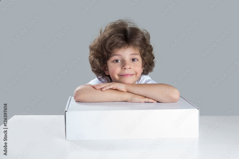 Cute boy smiling with his head resting on his folded hands on the white box. He sits at a table looking at the camera. Gray background. Close-up. Cute boy smiling with his head resting on his folded hands on the white box. He sits at a table looking at the camera. Gray background. Close-up.