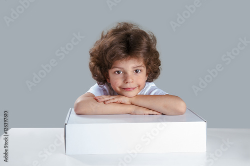 Cute curly little boy sitting at a table with her hands folded on a white box. He looks into the camera. Gray background. Close-up.