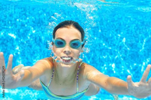 Child swims in pool underwater, funny happy girl in goggles has fun under water and makes bubbles, kid sport on family vacation 