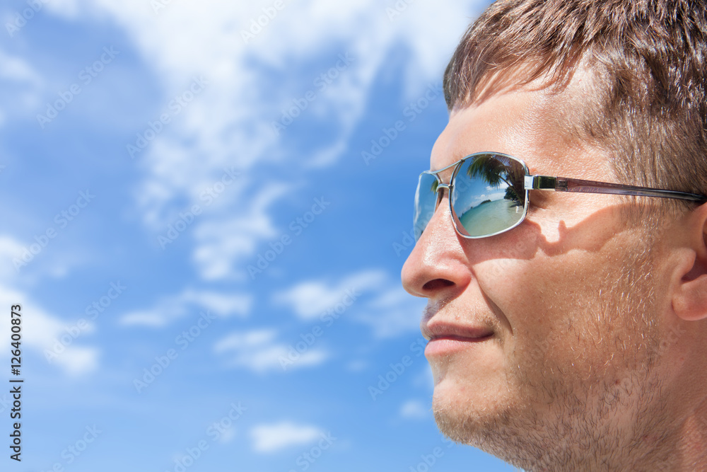 Young man looks at an exotic beach on blue sky with clouds background, reflection in sunglasses. Close up portrait with copy space Young man looks at an exotic beach on blue sky with clouds background, reflection in sunglasses. Close up portrait with copy space