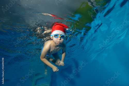 Child in hat Santa Claus swims underwater in the pool on a blue background and looks at me. The view from under the water at the bottom. Horizontal view