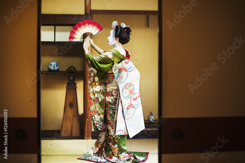 A woman dressed in the traditional geisha style, wearing a kimono and obi, with an elaborate hairstyle and floral hair clips, with white face makeup with bright red lips and dark eyes. Standing in a classic pose with fan raised, side view. 
