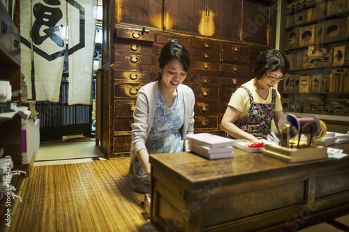 A traditional wagashi sweet shop. A woman working at a desk using a laptop and phone. A woman packing merchandise. 
