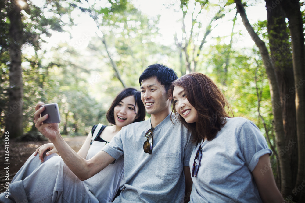 Two young women and a man sitting in a forest, taking a selfie. Two young women and a man sitting in a forest, taking a selfie.