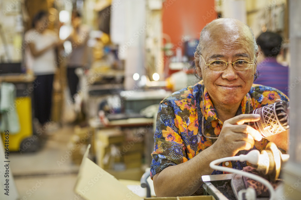 A senior craftsman in a glass maker's workshop sitting at a workbench using a light to check an etched or cut glass pattern. A senior craftsman in a glass maker's workshop sitting at a workbench using a light to check an etched or cut glass pattern.