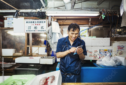 A traditional fresh fish market in Tokyo. A man in a blue apron standing behind the counter of his stall, using a smart phone covered with protective plastic.  photo