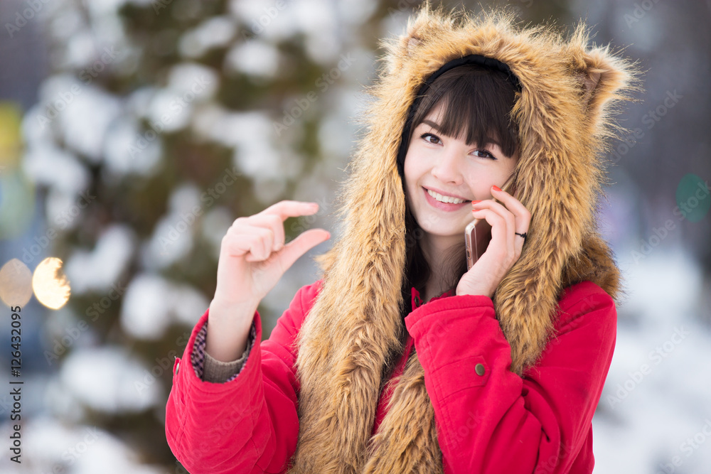 Portrait of smiling beautiful young woman in red winter coat and funny fur hat posing with phone, making a call outdoors in wintertime, showing something invisible and small in hands Portrait of smiling beautiful young woman in red winter coat and funny fur hat posing with phone, making a call outdoors in wintertime, showing something invisible and small in hands