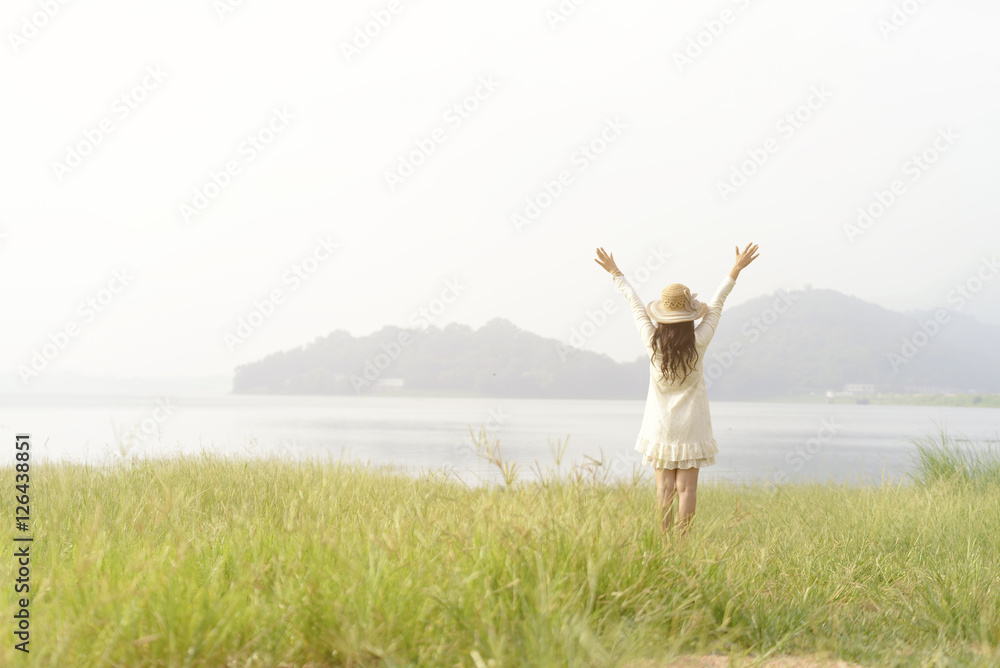 A pretty woman wearing white dress standing arms outstretched in the middle of the grass with reservoir and mountain background. A pretty woman wearing white dress standing arms outstretched in the middle of the grass with reservoir and mountain background.