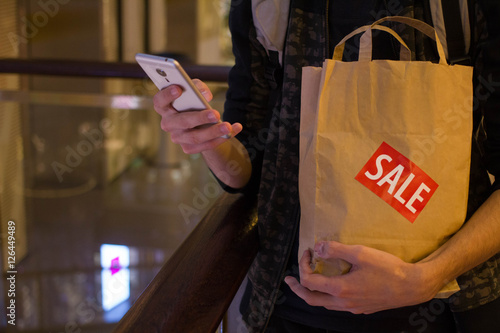 Man hold brown paper bag with SALE sticker on it in one hand and use smartphone with another in mall photo