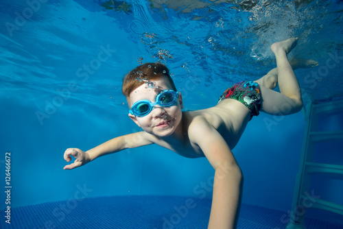 Little boy swims underwater in the pool, smiling, blowing bubbles and looking at me. The view from under the water. Close-up. Horizontal orientation