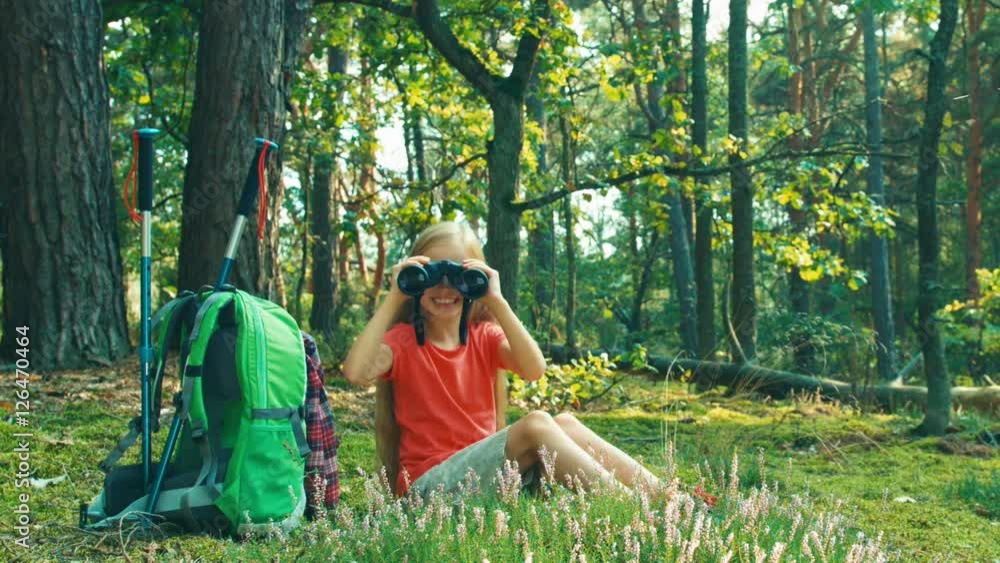 Hiker girl 8-9 years child using binoculars and looking at camera in the forest. Child waving hand and smiling. Zooming Hiker girl 8-9 years child using binoculars and looking at camera in the forest. Child waving hand and smiling. Zooming