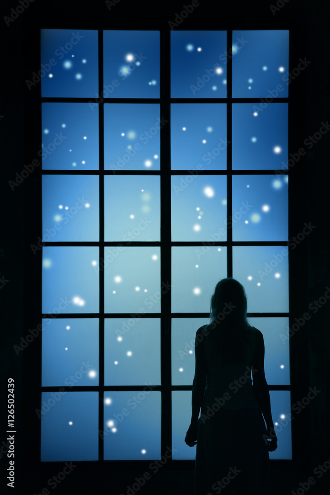 Woman silhouette watching the snowfall through big closed glass window and thinking about Christmas and New year things.