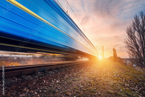 High speed blue passenger train on railroad track in motion at sunset. Blurred commuter train. Railway station. Railroad travel, railway tourism. Industrial landscape in the evening in autumn. Concept