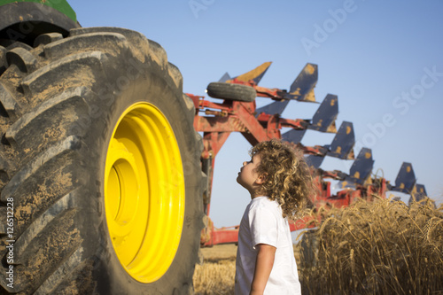 A Toddler looks up at the wheel of a big tractor with plough on the back in a wheat field photo