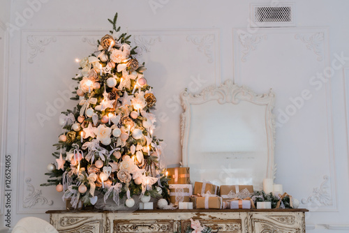 Christmas tree on wooden chest of drawers commode bureau in white interior, decorated with artificial flowers, garlands and toys