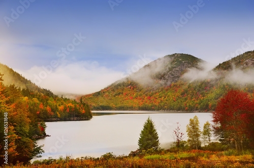 Lake in the mountains  colors around autumn trees.   Acadia National Park. Maine. USA. Cloudy gloomy weather  fog and clouds over the mountains in the distance.   