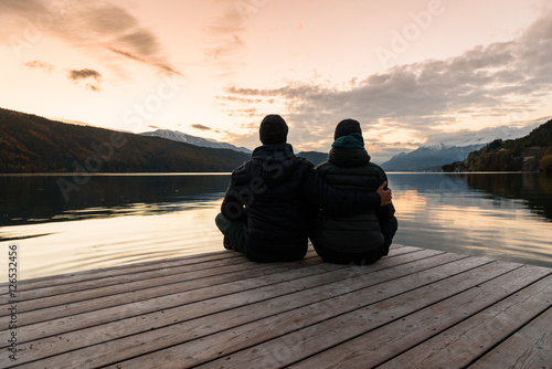 Rear view of a romantic young couple sitting on the pier enjoying beautiful sunset at lake. Autumn warm clothes. Love and meditation. 