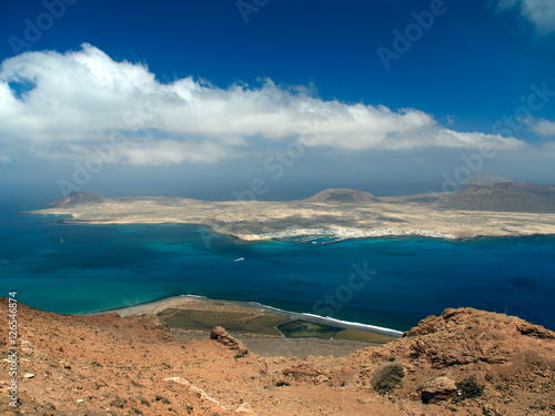 Sandy island with volcanoes and mountains in Atlantic Ocean. Aerial view from mountain slope. Yacht harbor and boats in strait. Deep blue sky with white clouds. La Graciosa, Canary Islands, Spain