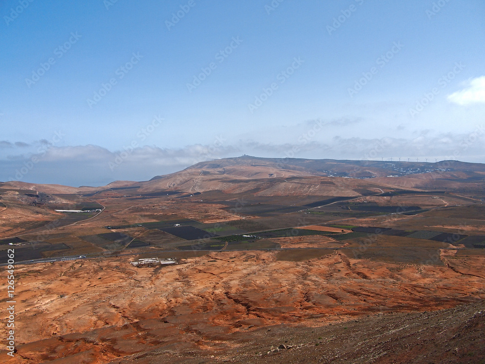 Mountain valley with white buildings and villages between old volcanic slopes. Red Earth and green fields on the background of deep blue sky with white clouds. Lanzarote, Canary Islands, Spain