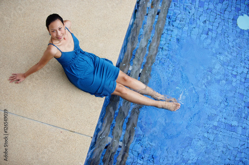 Young woman sitting by swimming pool, feet in water, smiling at camera