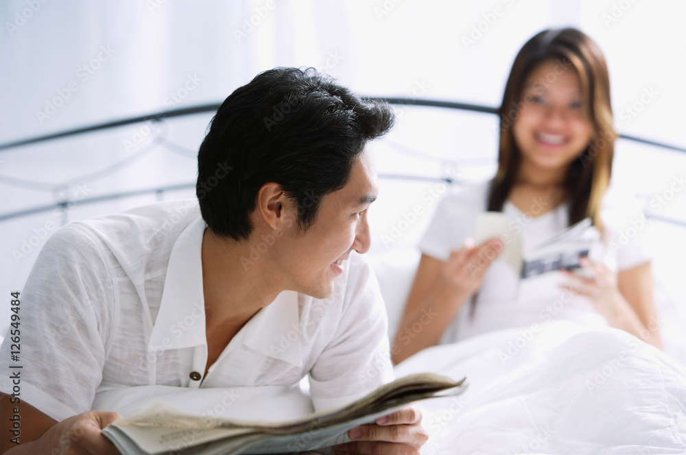 Couple on bed, man with newspaper, smiling at woman behind him