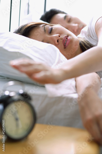 Couple lying on bed, woman reaching to switch of alarm clock in foreground